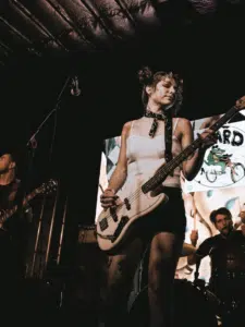 A woman plays an electric bass guitar on stage with a band at festivals, while a drummer and another guitarist perform in the background.